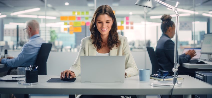 Young Happy Businesswoman Using Computer in Modern Office with Colleagues. Stylish Beautiful Manager Smiling, Working on Financial and Marketing Projects. Drinking Tea or Coffee from a Mug.