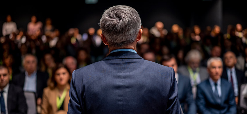 Back of middle aged man wearing dark gray suit looking at crowd who are looking back at him
