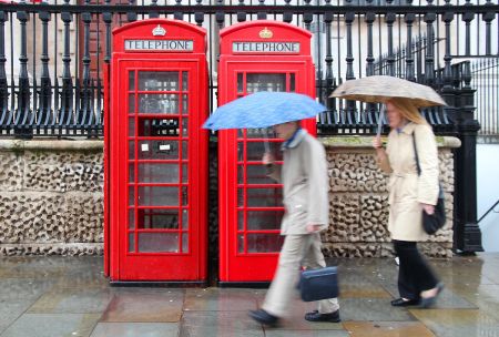 Two people holding umbrellas walk past typical red telephone booth in London, UK