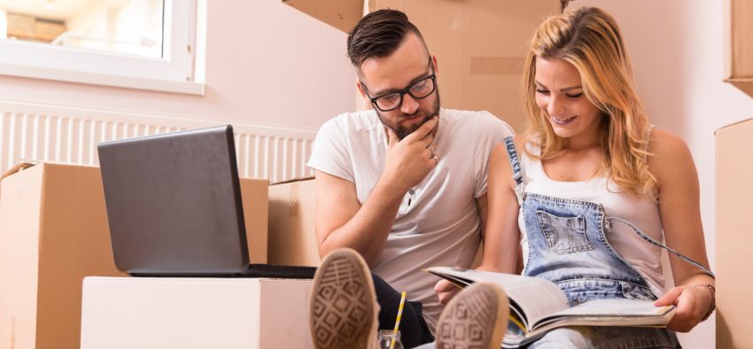 Young couple in love sitting on the floor of their new apartment, planning redecoration and searching for ideas on a laptop computer and in a home decorating magazine