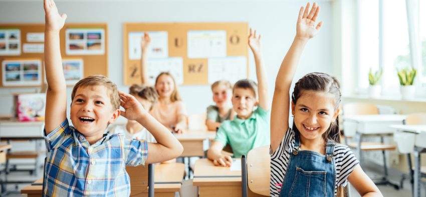 School children in classroom at lesson. Little children raising hands up and having fun in class.