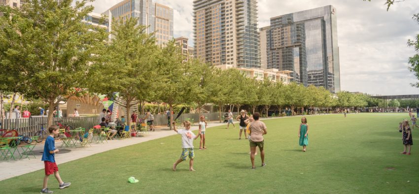 DALLAS, TX, USA-MAY 26, 2018:Klyde Warren Park, a 5.2-acre public park in downtown Dallas, Texas. People playing sport on green lawn grass under sunny day cloud blue sky. Live oak tree and skyscraper