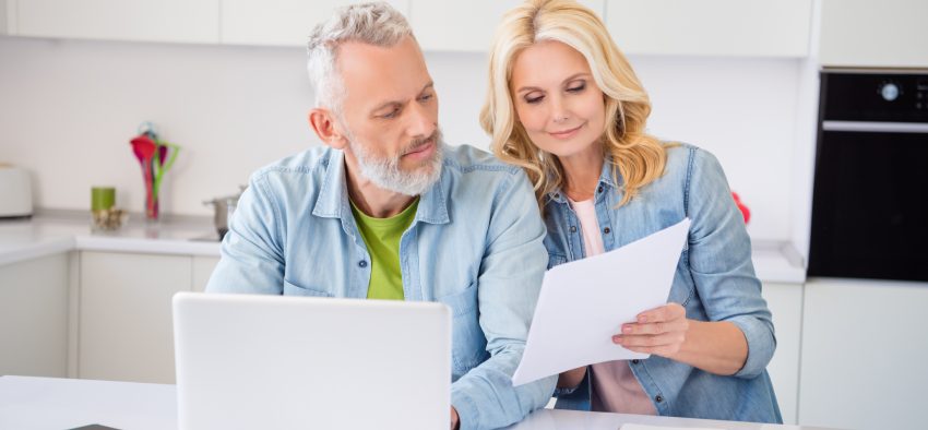 Photo of thoughtful pretty aged couple wear denim shirt smiling reading dicuments browsing modern device indoors house room