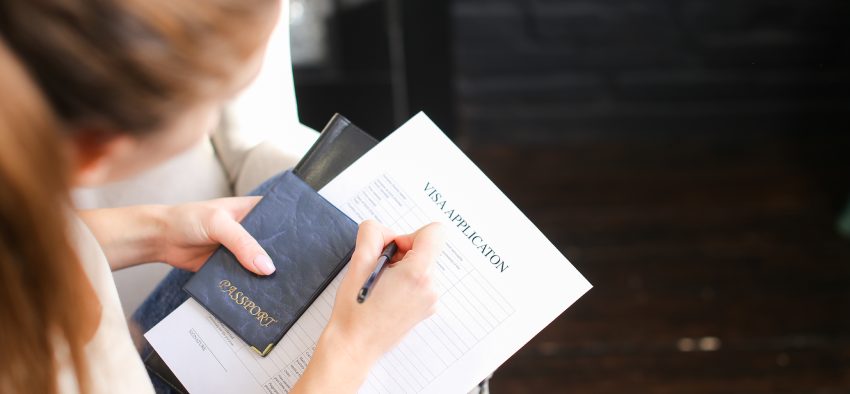 girl filling visa application form, student sitting on grey chair with passport. Young fair-haired woman wearing beige T-shirt jeans holding planchette with papers writing. Concept of execution of