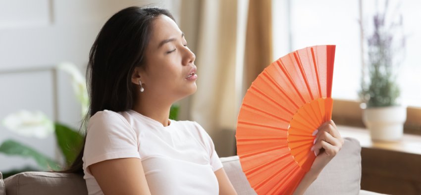 Overheated asian woman sweating feels discomfort seated on sofa at home without air-conditioning system. Tired girl suffers from heat stroke waving orange color fan cools herself in hot summer weather