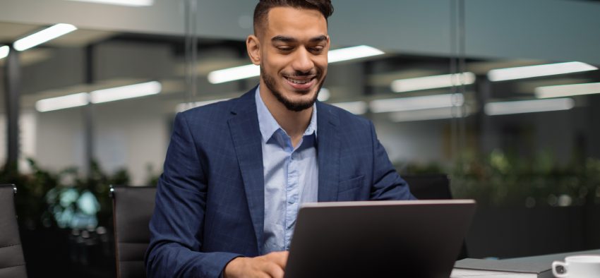 Smiling young middle eastern businessman sitting at worktable at modern office, typing on computer keyboard, sending emails to his business partners, working on marketing research, copy space