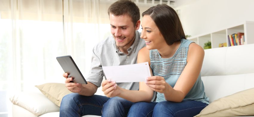 Happy couple checking bank account on line in a tablet sitting on a couch in the living room at home