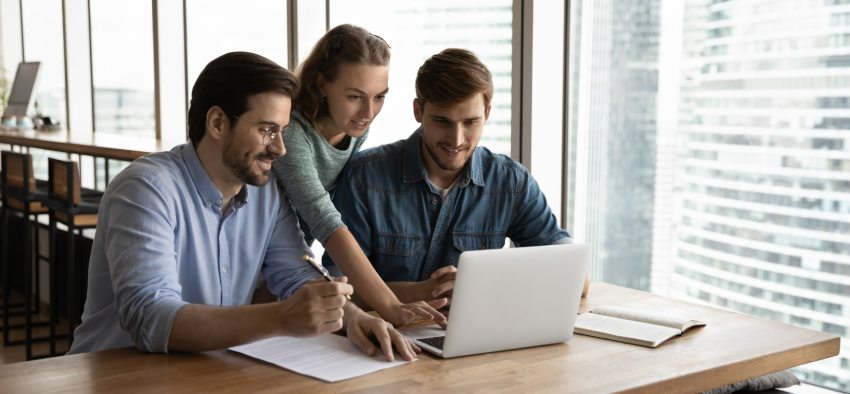 Creative business team sharing laptop in office, watching startup presentation, design project, discussing ideas, brainstorming, pointing at monitor, talking. Office employees meeting at computer
