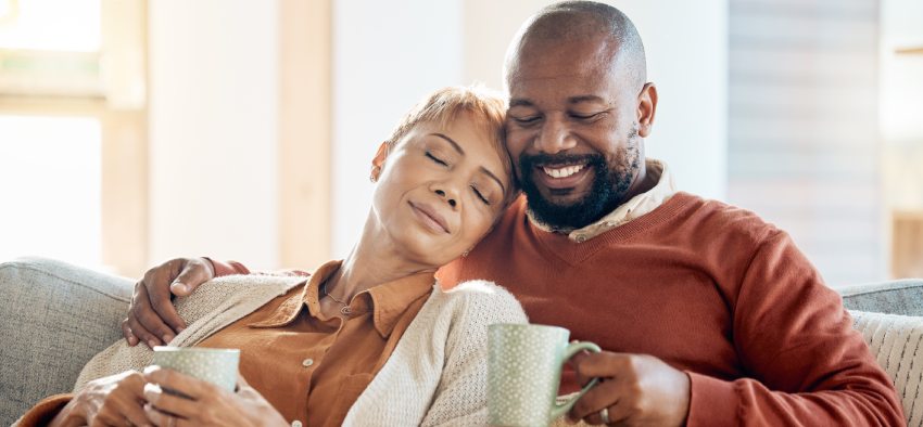 Black couple relax on sofa with coffee, blanket and smile on winter weekend morning in home. Peace, comfort and love, happy man and tired woman, cozy time on couch with drink in living room together.