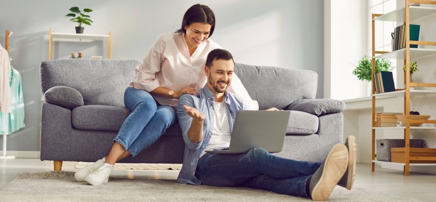 Happy couple having video chat using laptop computer at home. Smiling wife sitting on couch and hugging her husband who sitting on floor enjoying virtual meeting and talking. Online communication