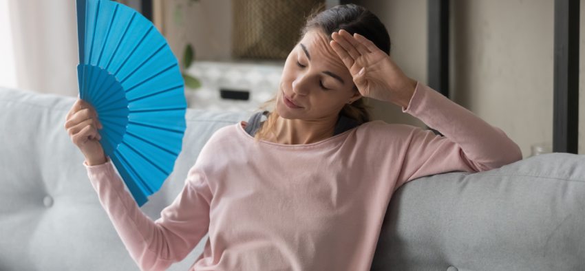 Overheated female sitting on couch in living room at hot summer weather day feeling discomfort suffers from heat waving blue fan to cool herself, girl sweating dwelling without air conditioner concept